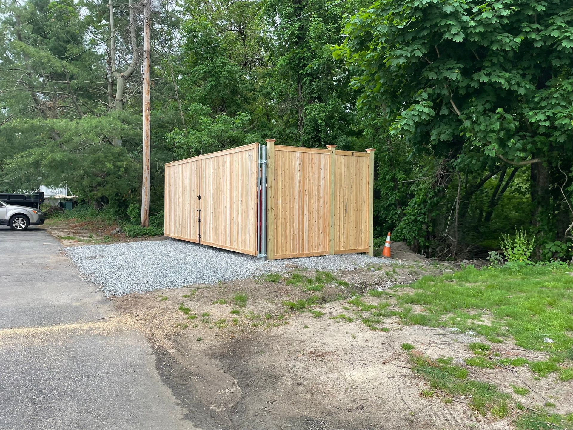 Wooden privacy fence enclosing a gravel area beside a wooded roadside, with an orange cone nearby.