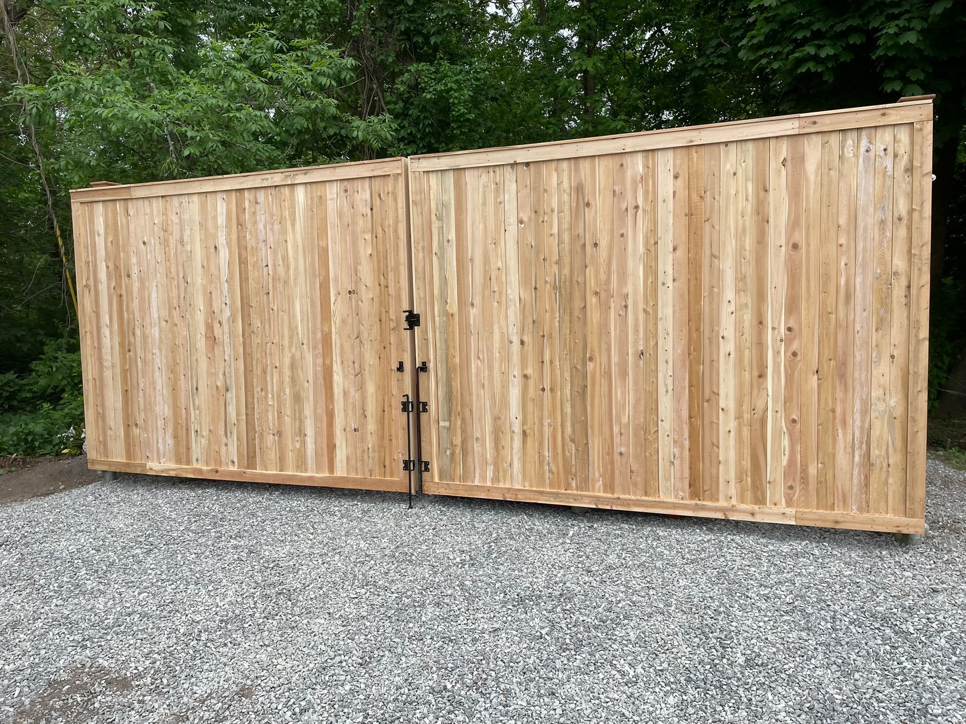 Wooden privacy fence with double gates on a gravel driveway, backed by green trees.