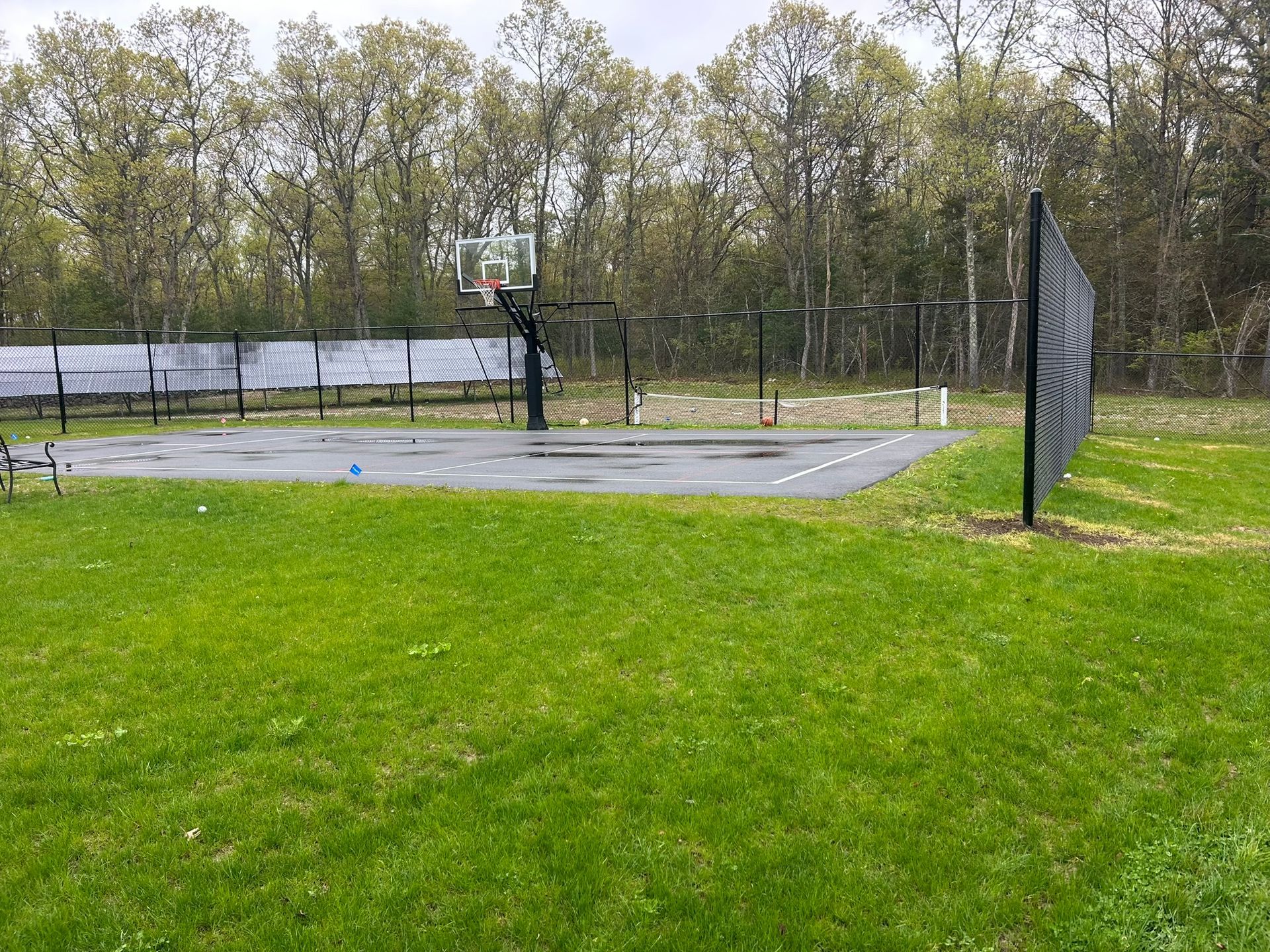Empty outdoor basketball court beside a grassy field and trees under an overcast sky