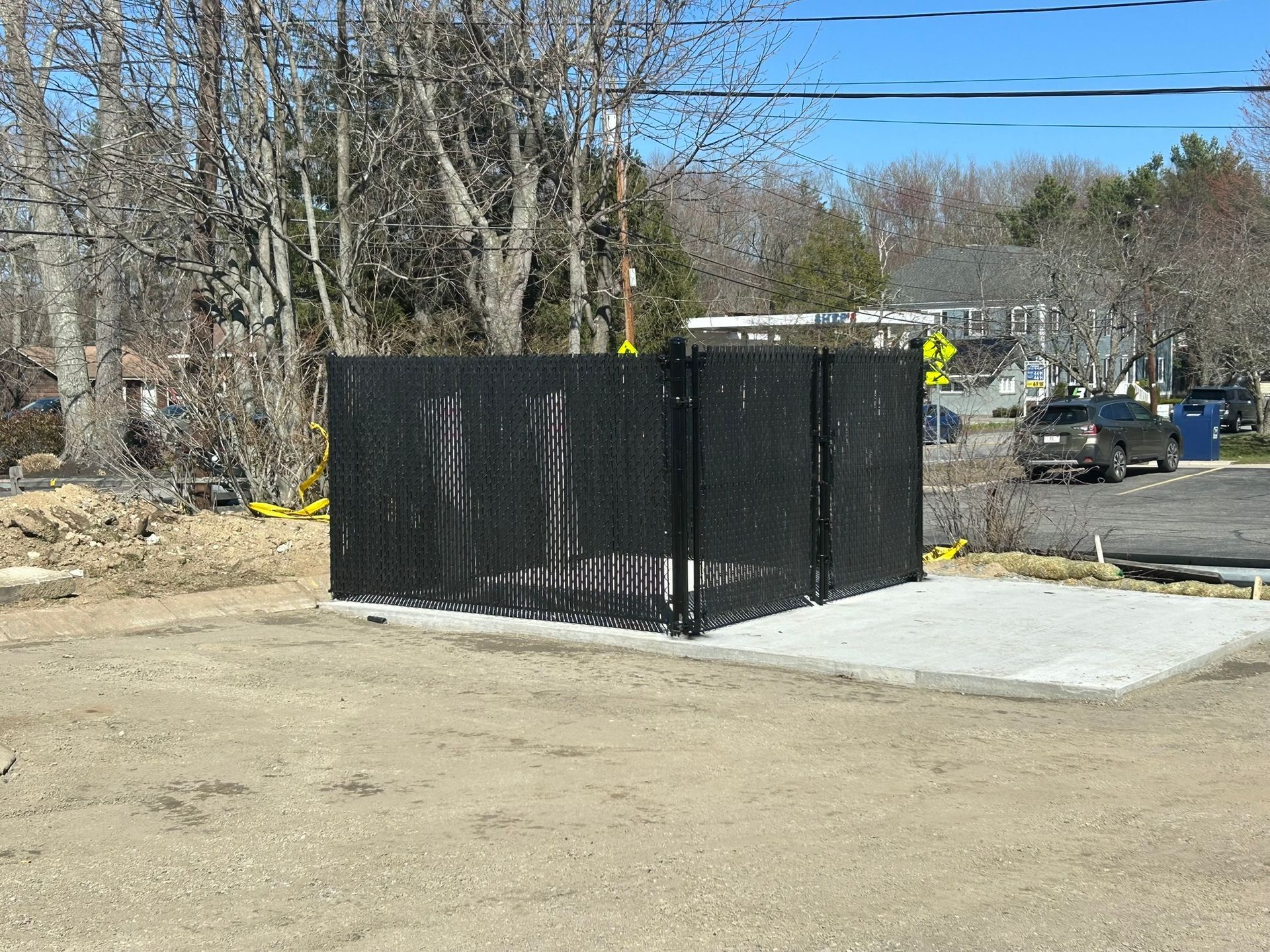 Black fenced enclosure on a concrete pad in a roadside outdoor area with trees and parked cars nearby