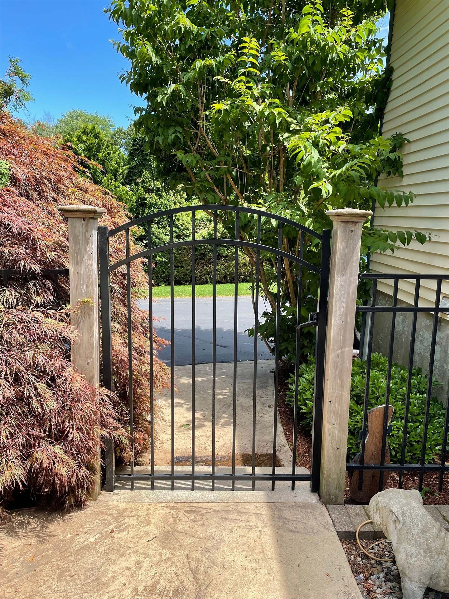 A wrought iron gate is leading to a driveway surrounded by trees and bushes.