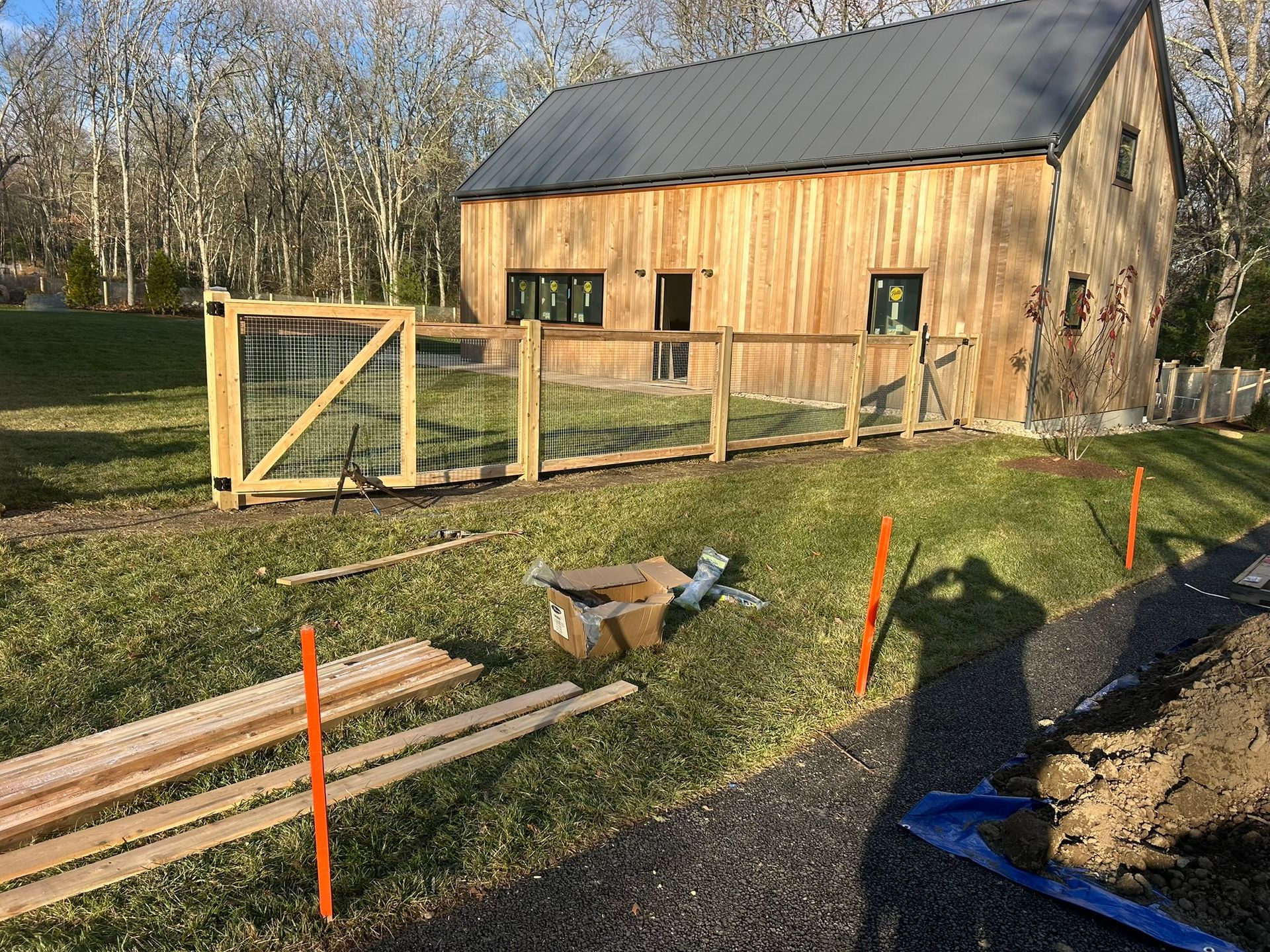 Wooden barn with fenced yard on a sunny grassy property, construction materials and orange stakes in foreground