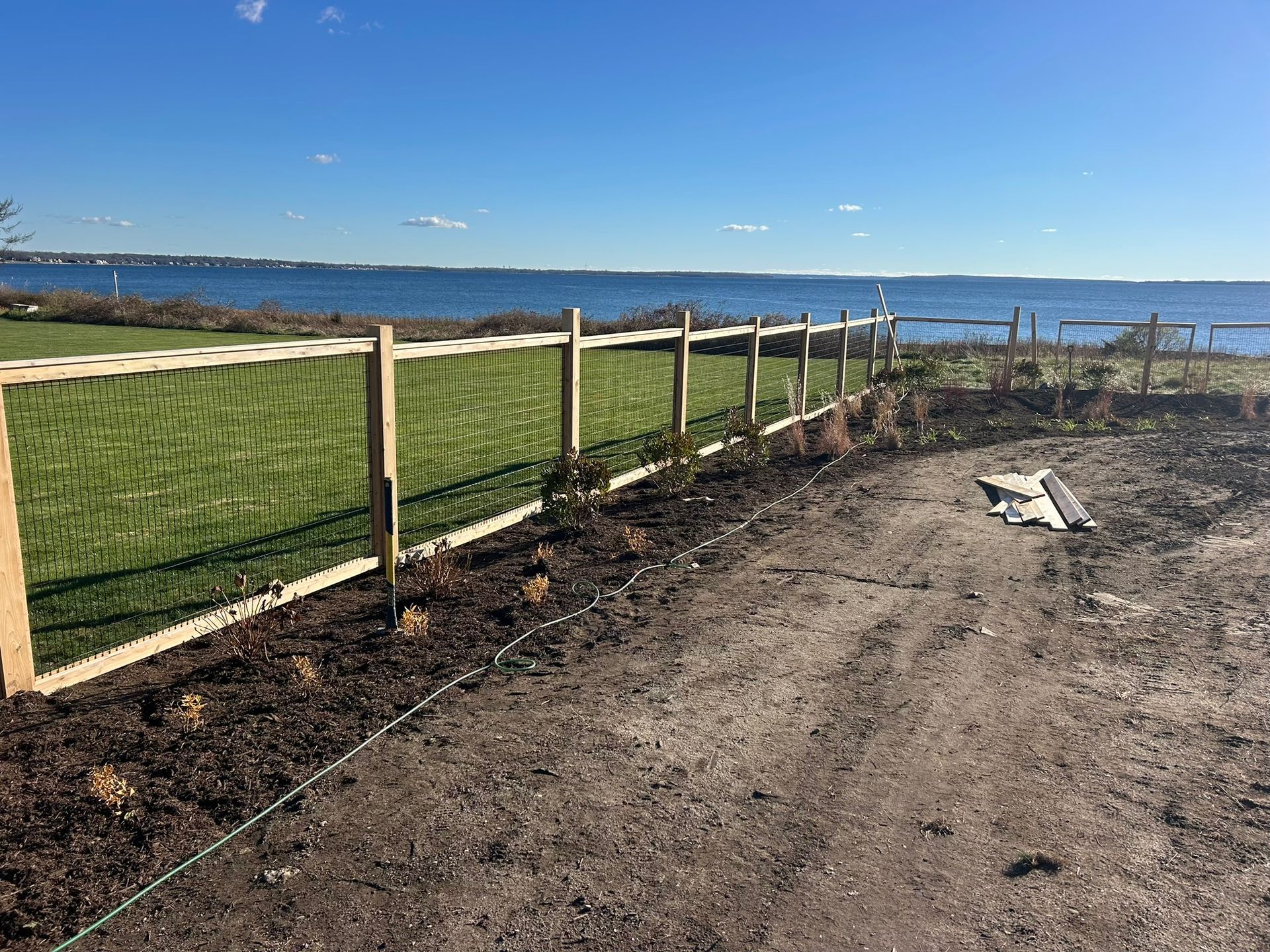 Fence line beside a dirt path overlooking a green field and calm blue water under a clear sky