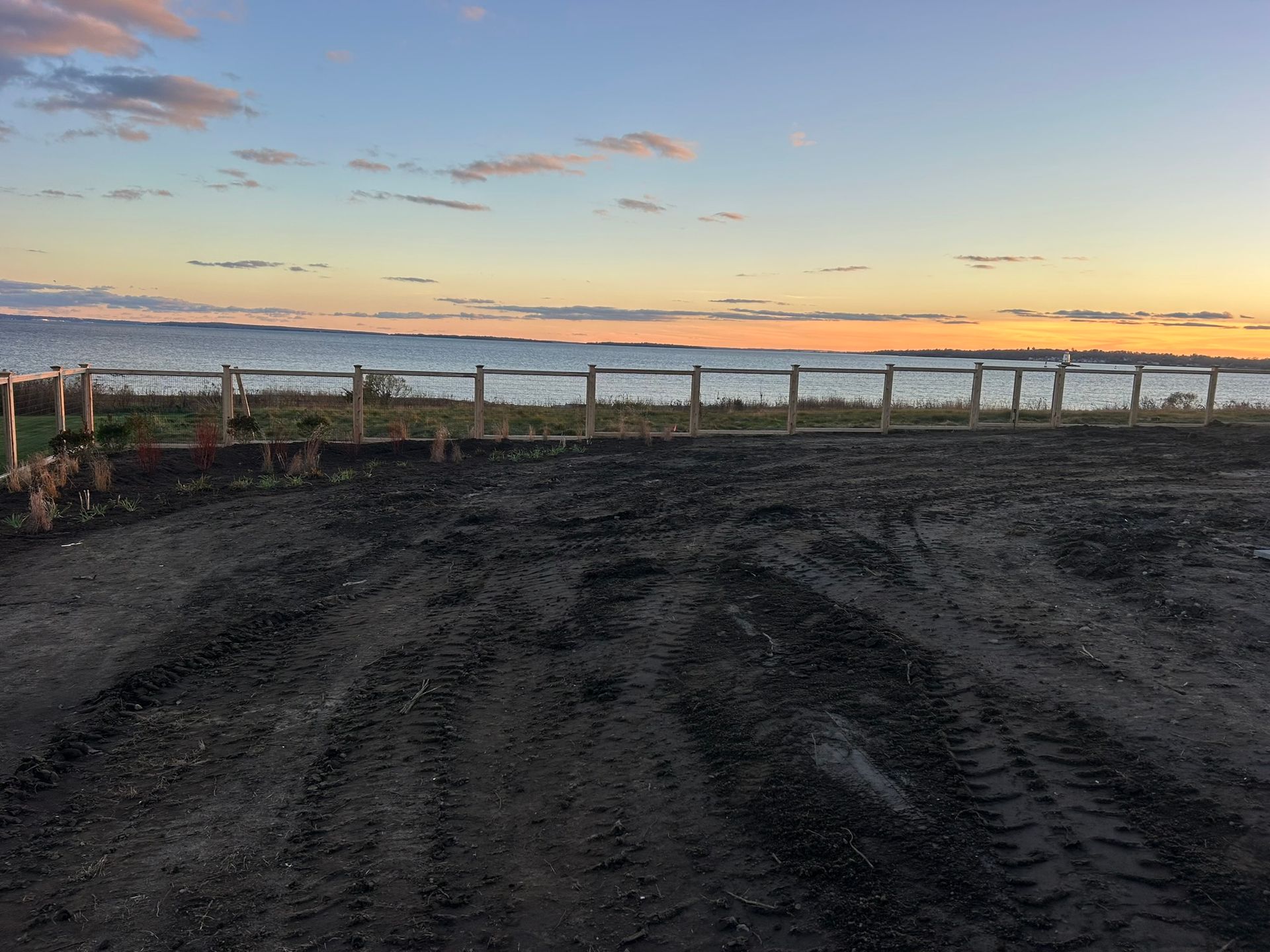 Gravel lot beside a waterfront at sunset, with a fence and calm sky in the distance