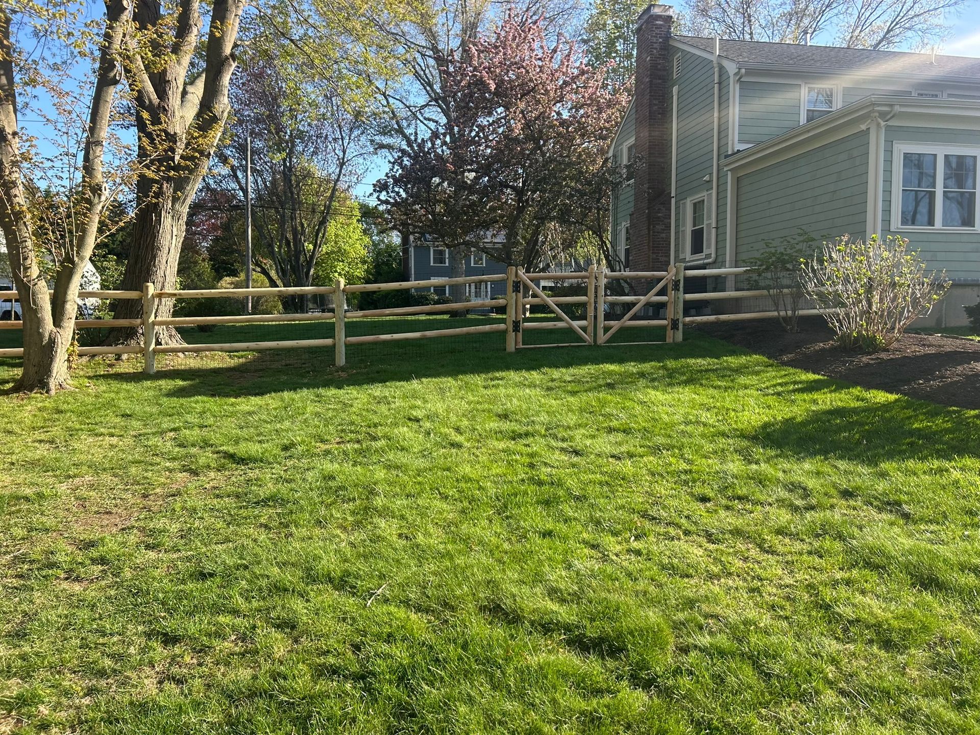 Sunny backyard with green lawn, wooden fence, trees, and a house deck with stairs