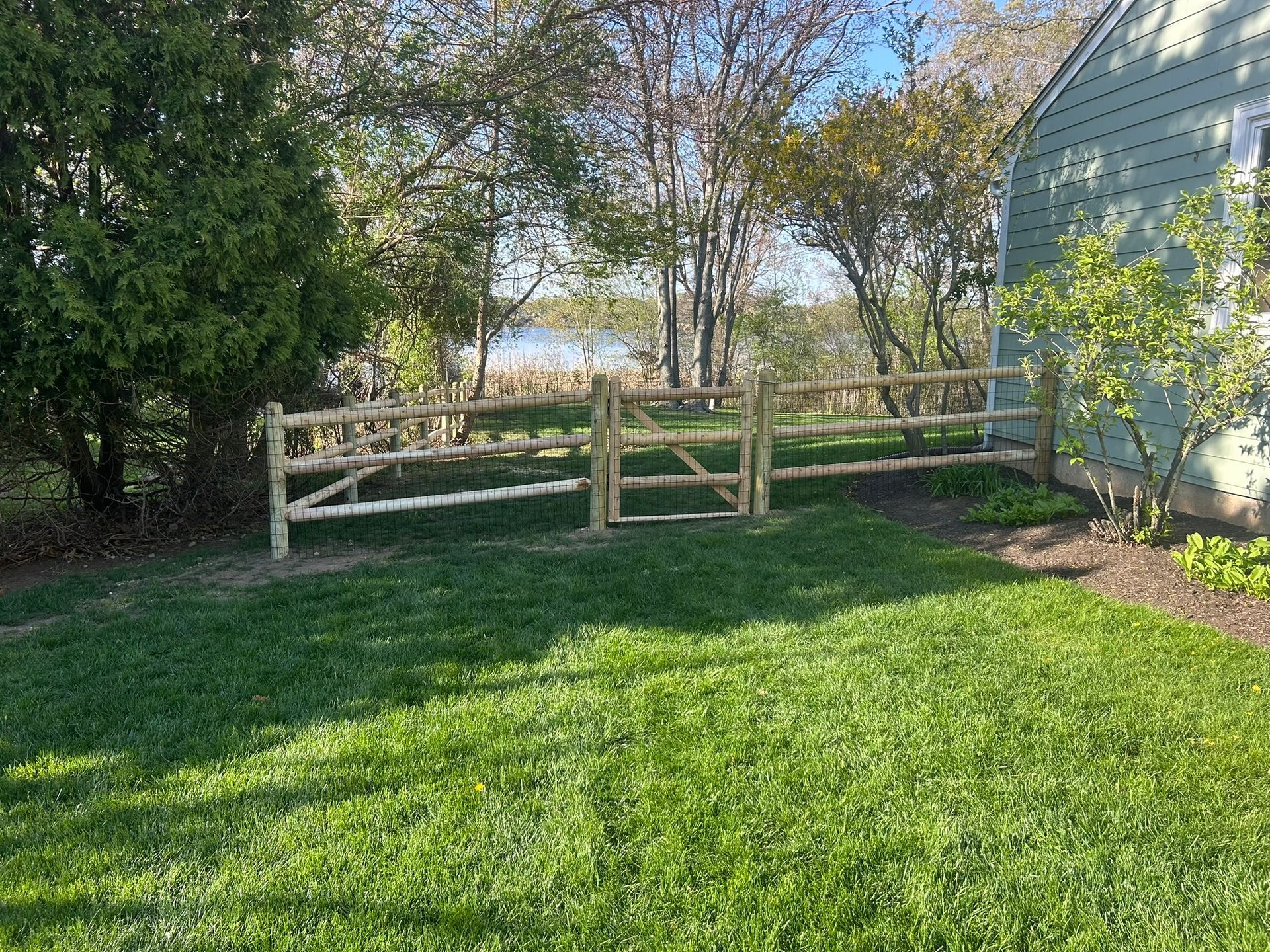 Wooden fence in a grassy backyard beside a house, with trees and sunlight.