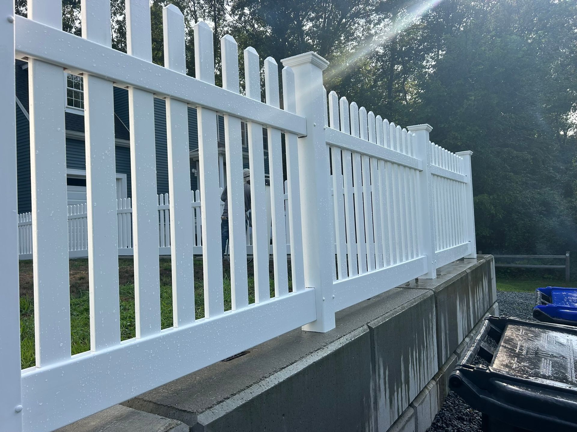White picket fence atop a concrete retaining wall beside a driveway, with trees in the background.