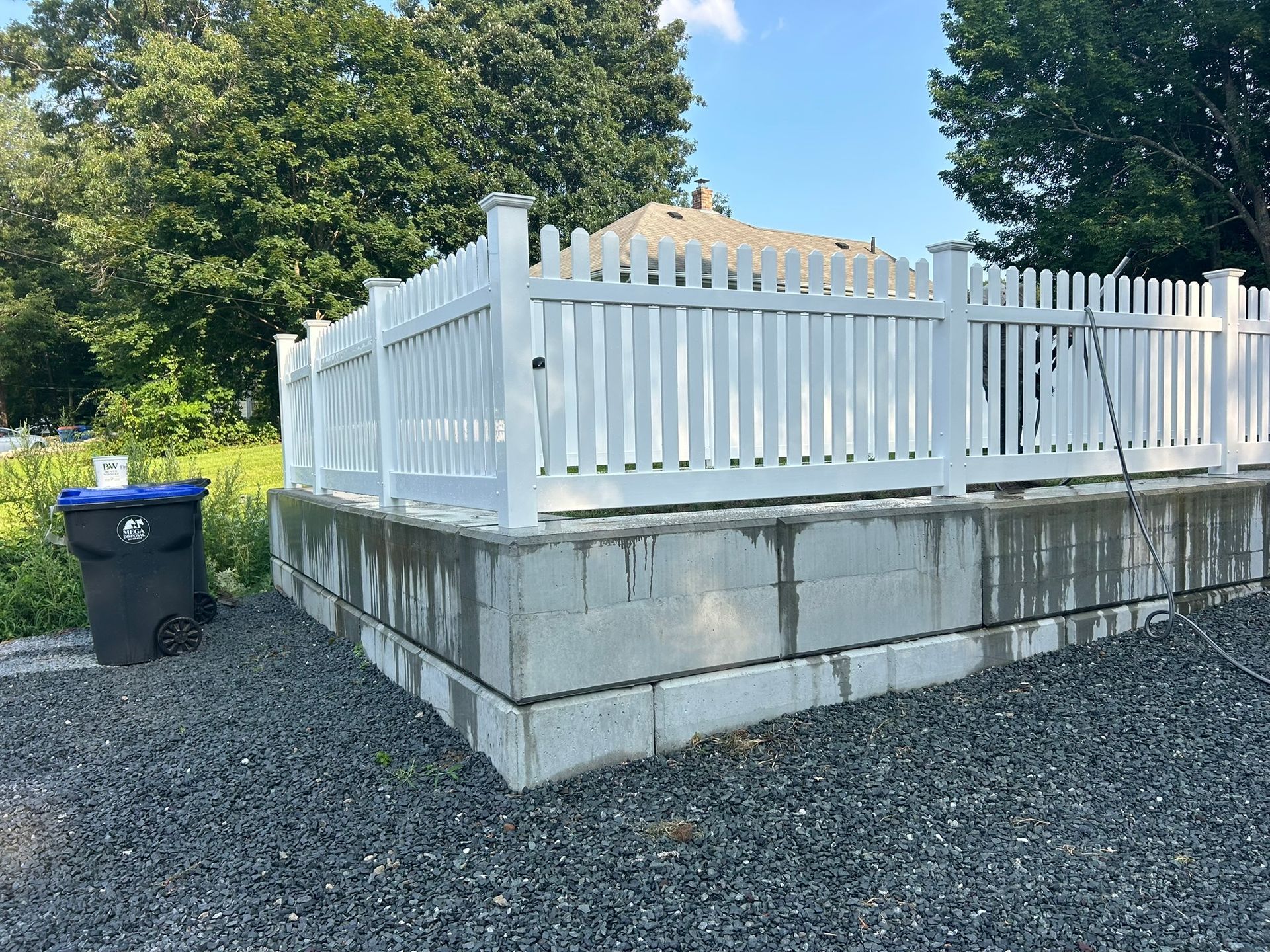 White picket fence atop a concrete retaining wall beside a gravel area and blue trash bin.