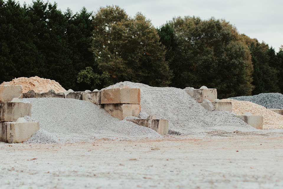 Piles of gravel, sand, and stone blocks sit in an outdoor yard, with trees in the background.