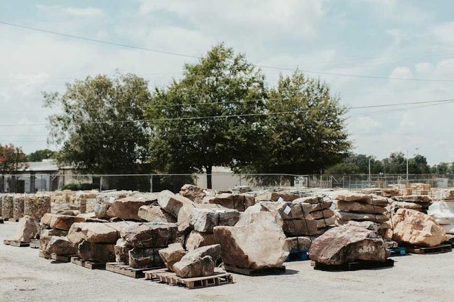 Rocks piled on pallets at an outdoor landscape supply store, trees in background, cloudy sky.