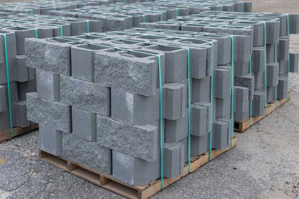 Stacks of gray concrete blocks on pallets, secured with blue straps, outdoors.