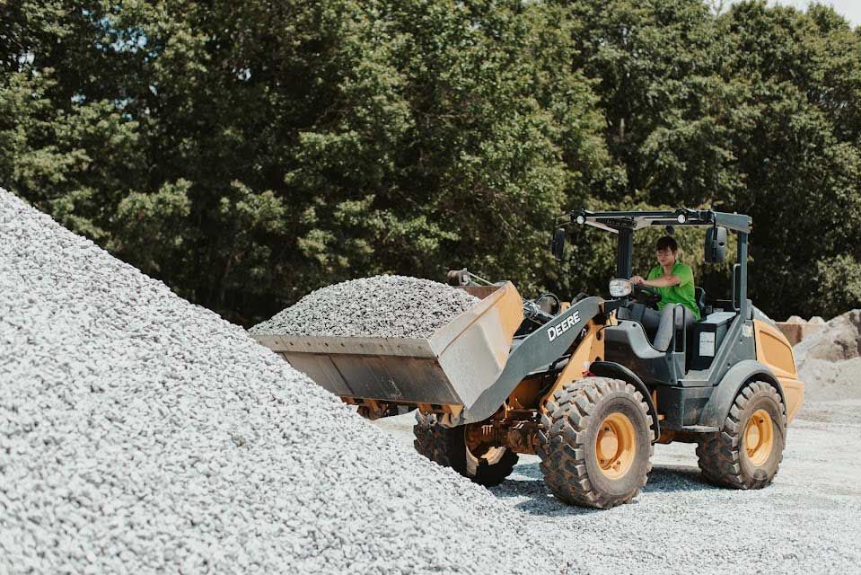 Loader with a person in the driver's seat scoops up gravel from a large pile, outdoors.
