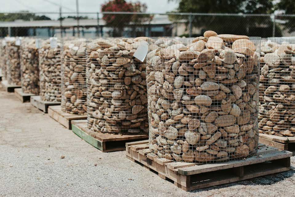Stone-filled wire baskets on pallets, likely for landscaping. Outdoor setting with fence and buildings.