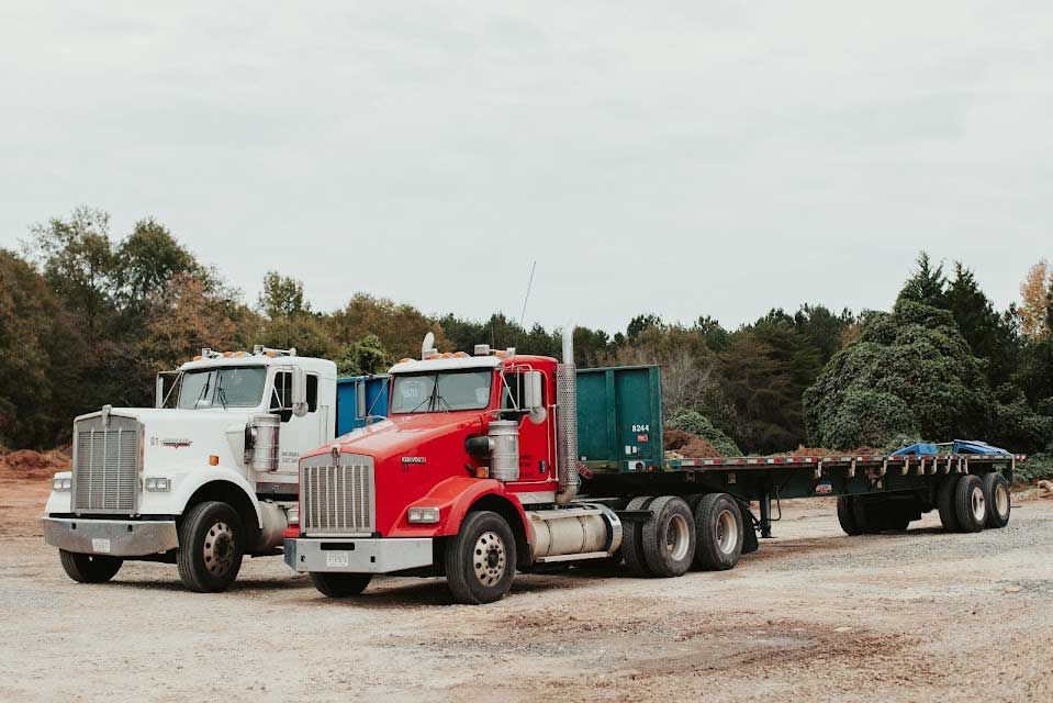Two semi-trucks parked on a gravel lot; white truck, red truck with flatbed trailer.