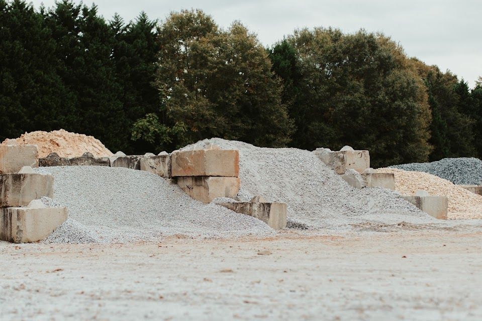 Piles of gravel and sand in various colors at a construction site with trees in the background.