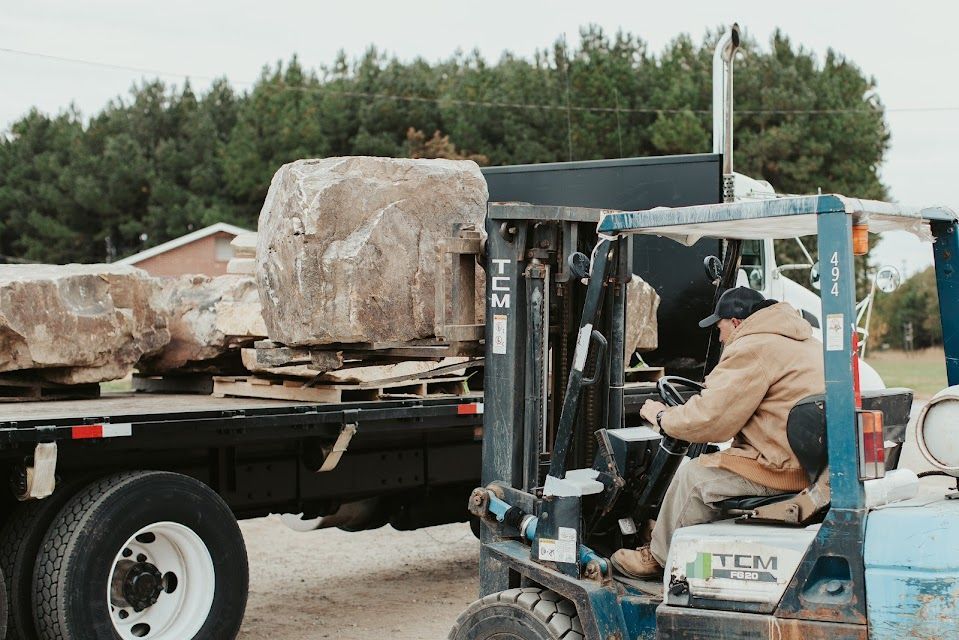 Forklift operator loading large rocks onto a flatbed truck outdoors.