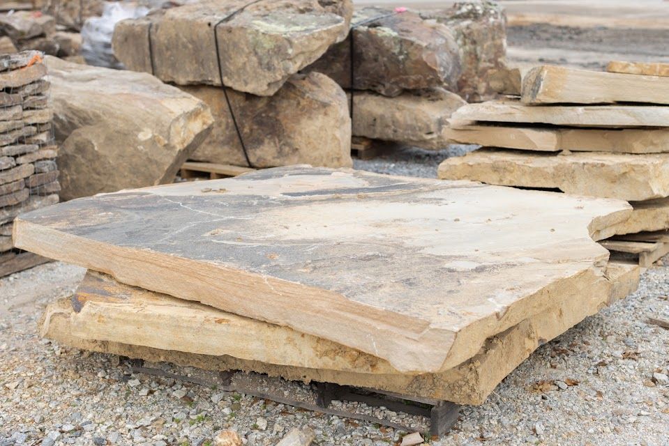Stacked, light-colored flagstones with rough edges. Other stone pieces and a pallet are in the background.