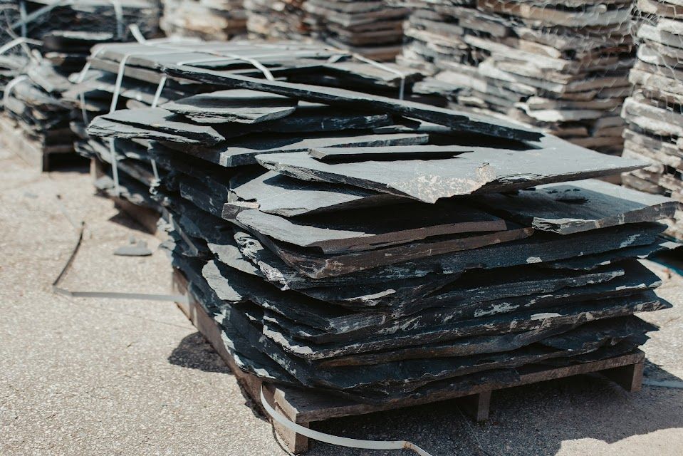Stacked slate tiles on wooden pallets, outdoors. Dark gray, irregular shapes.