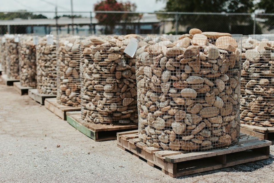 Rows of large wire baskets filled with beige river rocks on wooden pallets, outdoors.