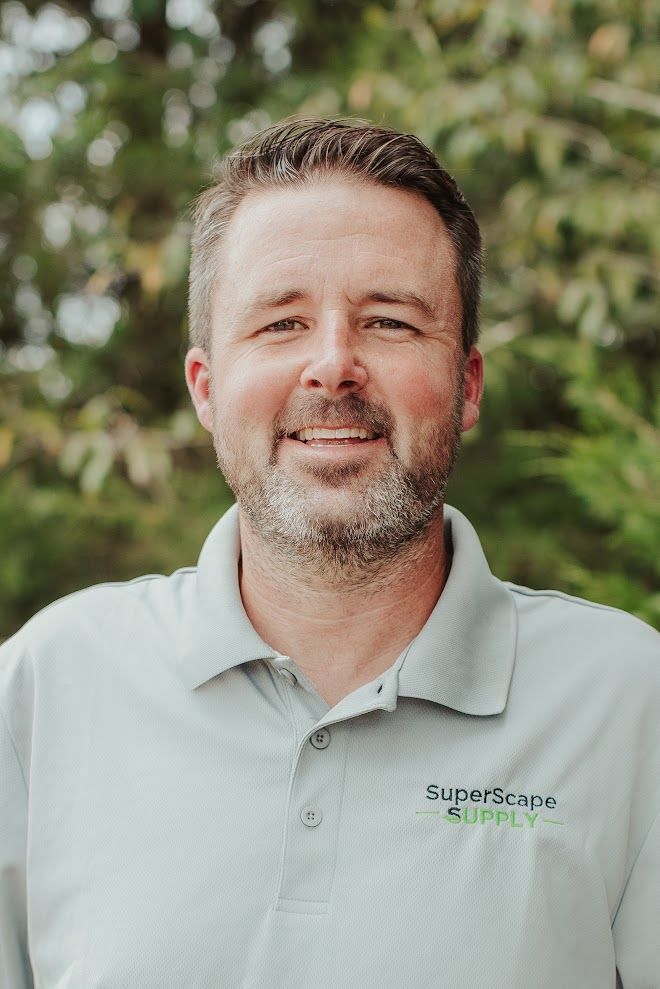 Man in grey polo shirt smiling, standing outdoors with a blurred green background. Shirt reads 