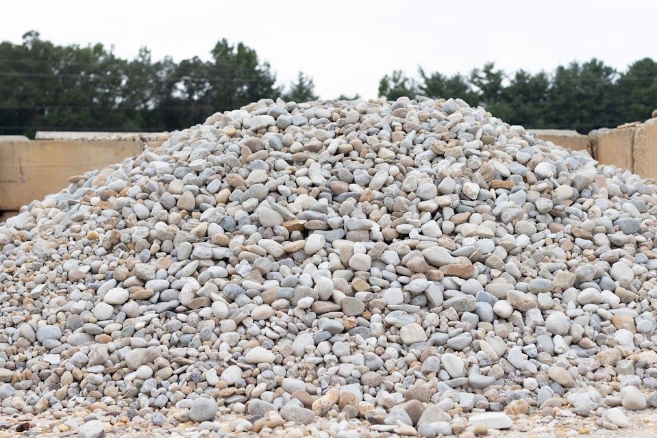 Pile of rounded, light-colored gravel against a background of green trees and concrete blocks.