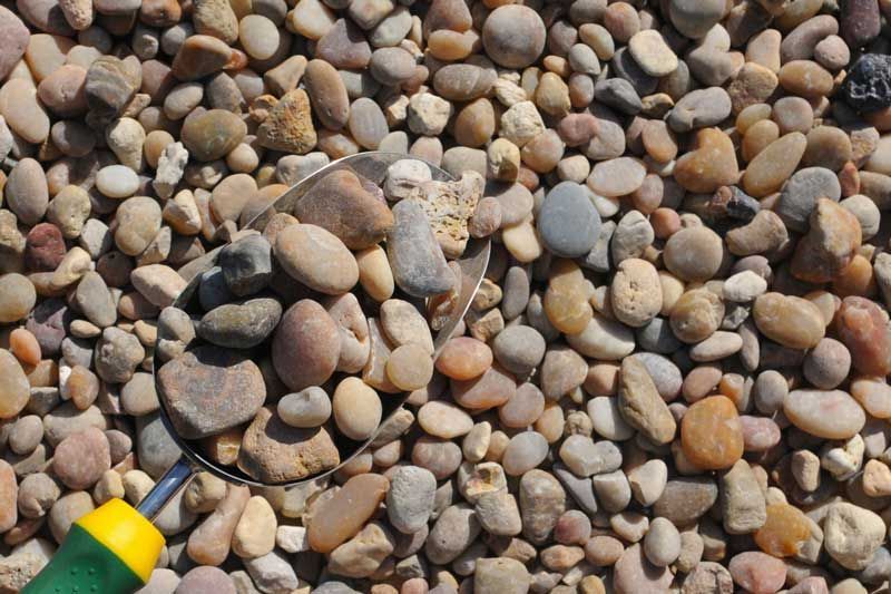 Small rocks in shades of brown and gray fill a metal scoop, reflecting sunlight.