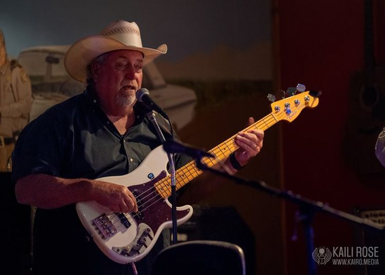 A man in a cowboy hat is playing a bass guitar in front of a microphone.