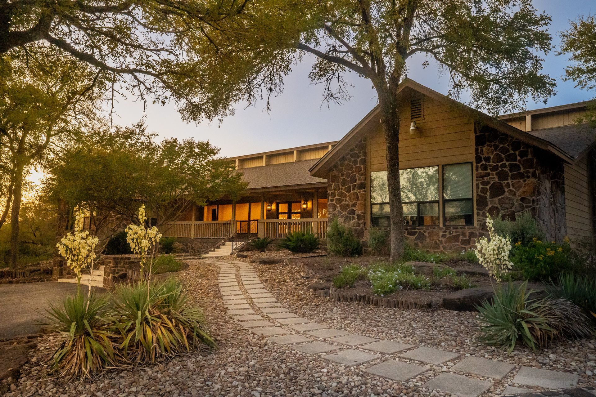 A large stone house with a stone walkway leading to it surrounded by trees and bushes.