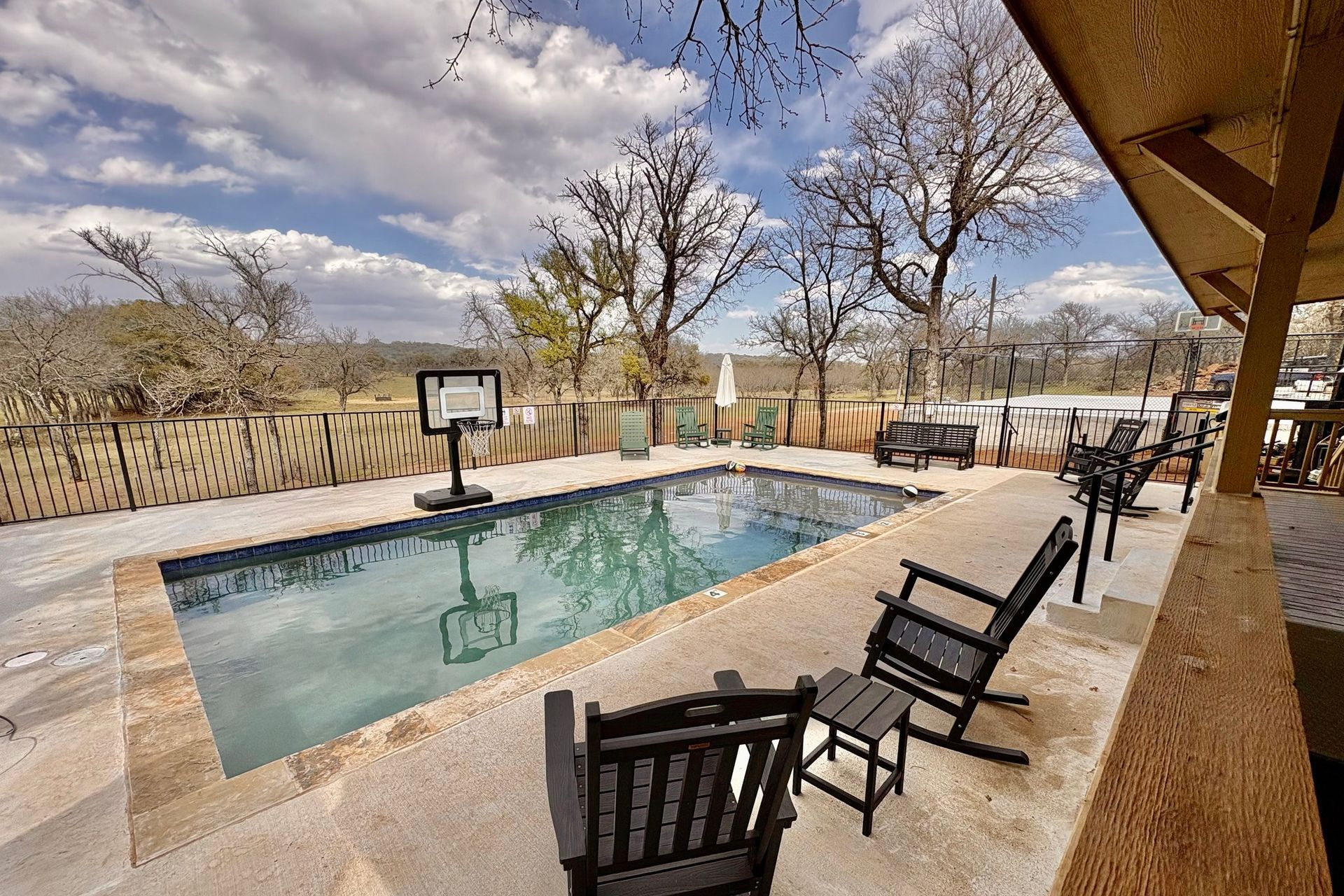 A large swimming pool is surrounded by rocking chairs and a basketball hoop.