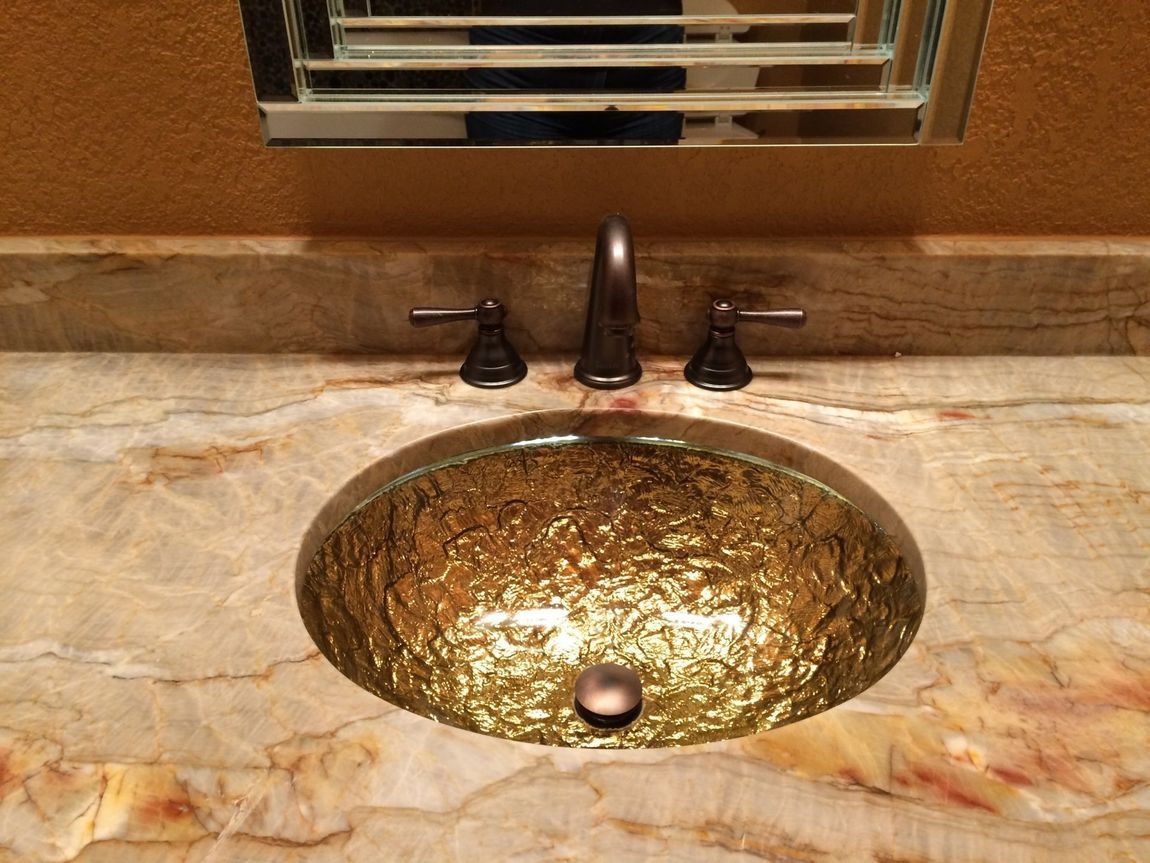 Gold textured glass sink on a marble countertop with dark bronze faucet and handles, reflected in a mirror.