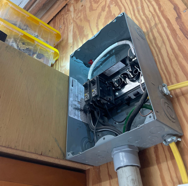 Open electrical panel with exposed wiring and breaker components mounted on a plywood wall.