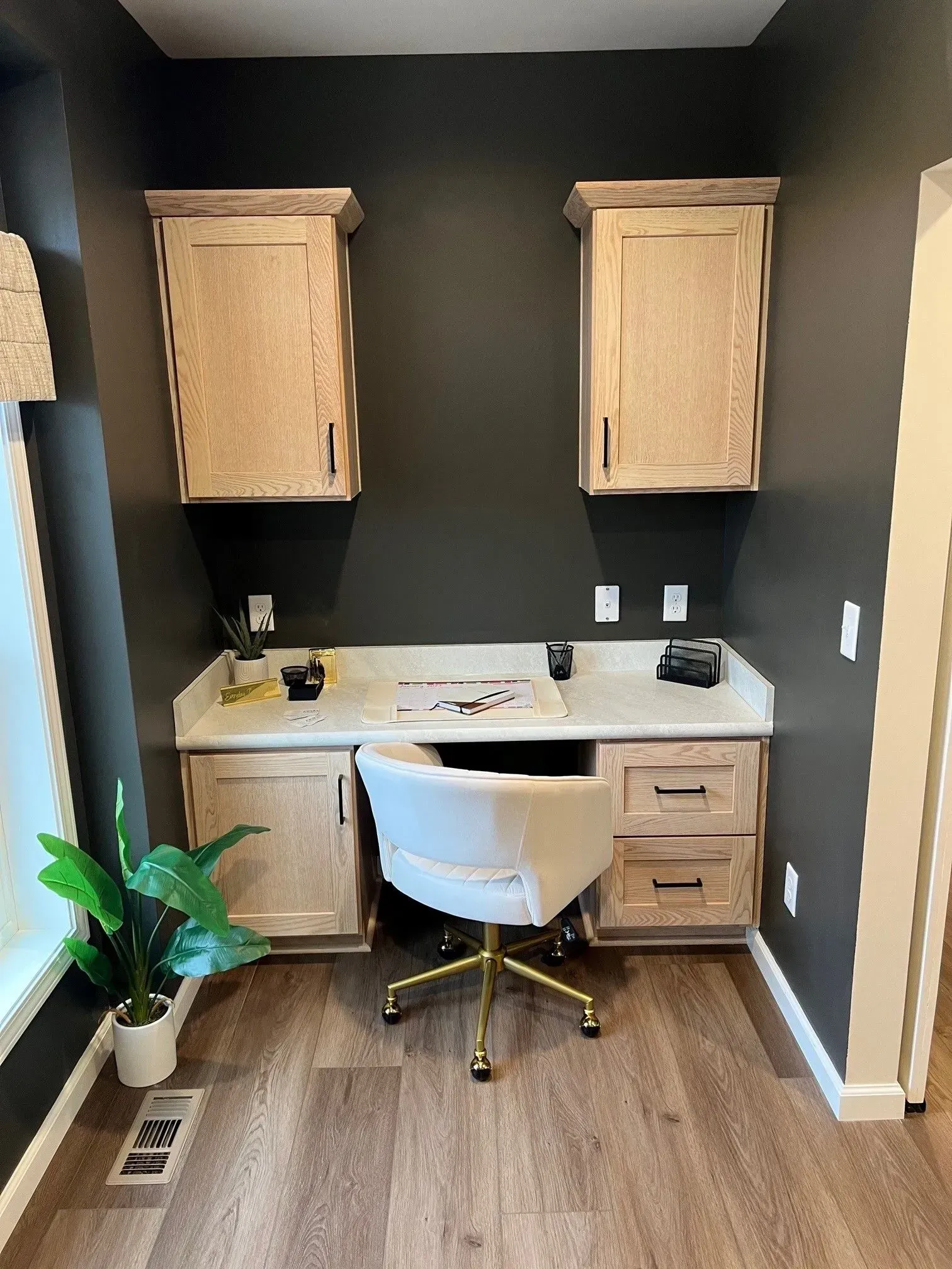 Home office with built-in desk, cabinets, and a gold-legged white chair. Dark gray wall, light wood cabinets/desk, light wood floor.