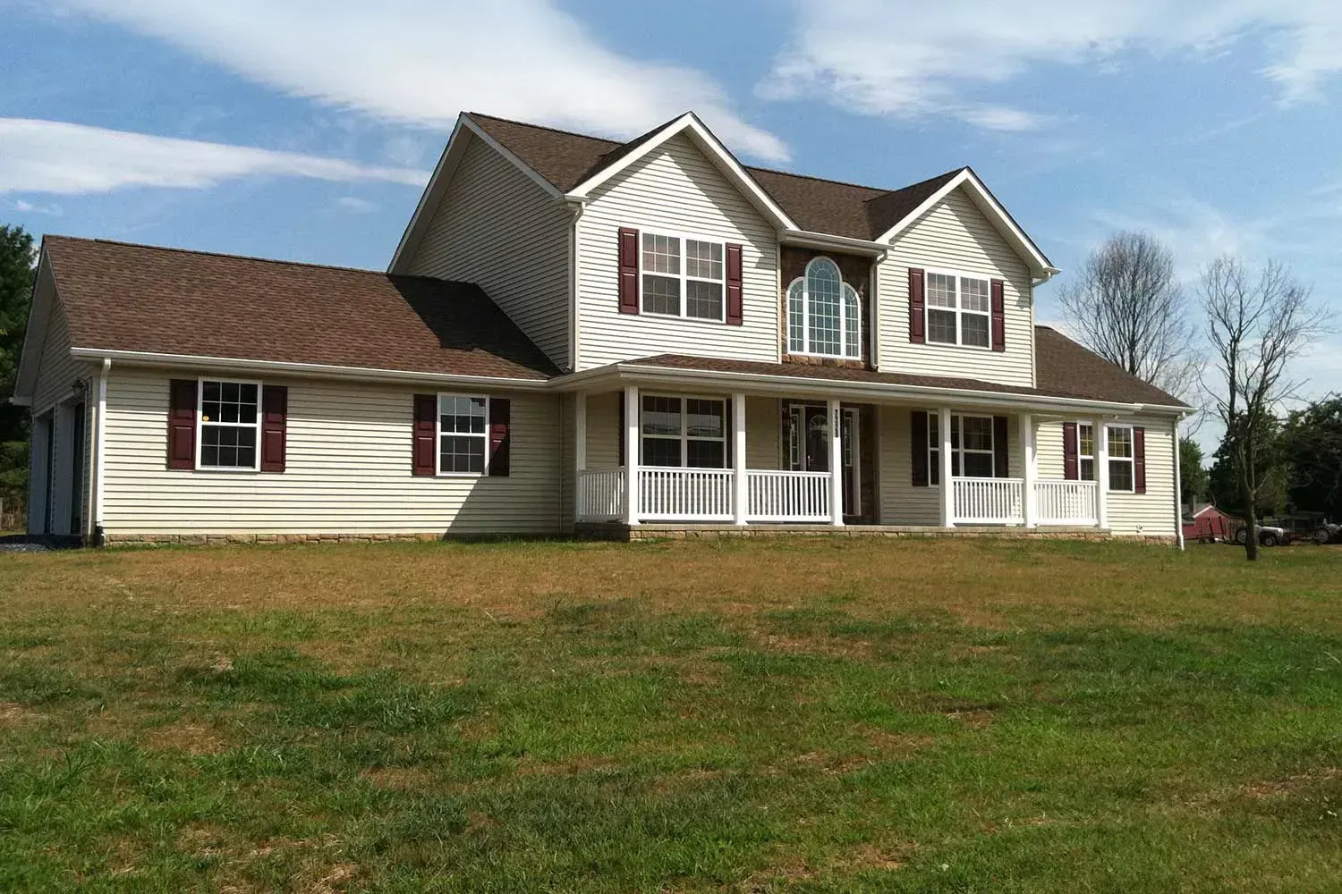 Two-story beige house with brown roof and burgundy shutters, porch, and grassy yard under blue sky.