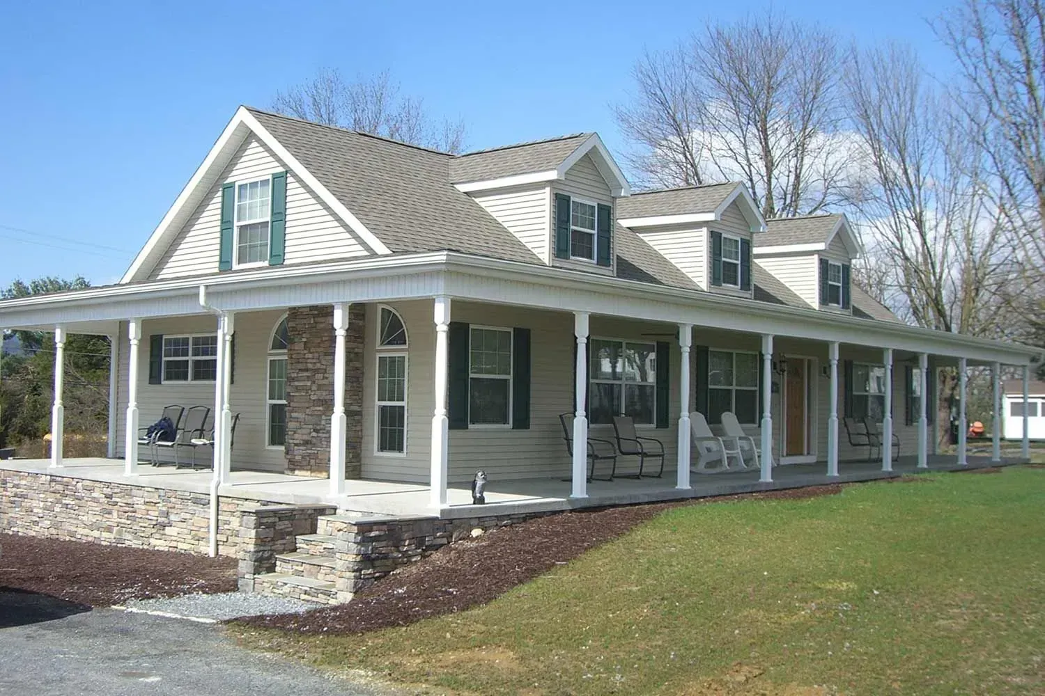 Tan house with a wraparound porch, green shutters, dormers, and a gray roof, in a grassy setting.