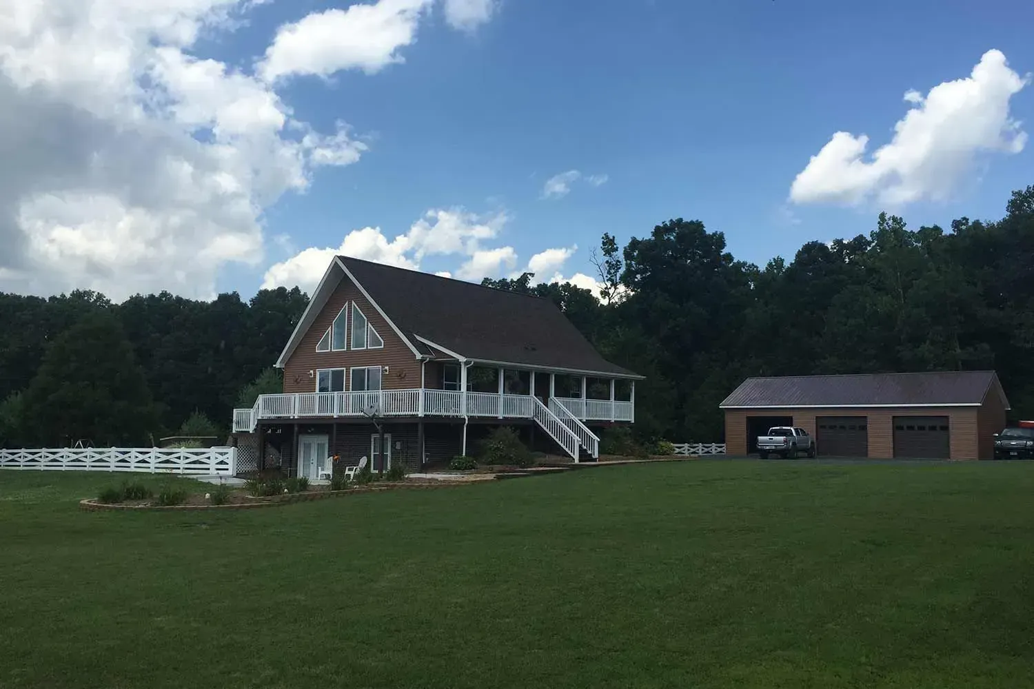House with wrap-around porch and attached garage on a grassy lot, trees in the background, blue sky with clouds.