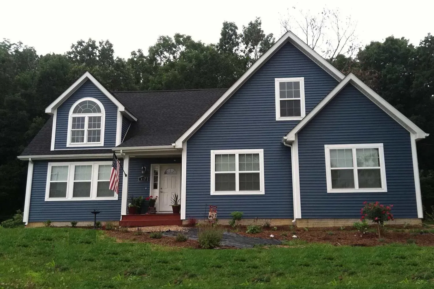 Blue-sided house with white trim, black roof, and manicured lawn in front of trees.