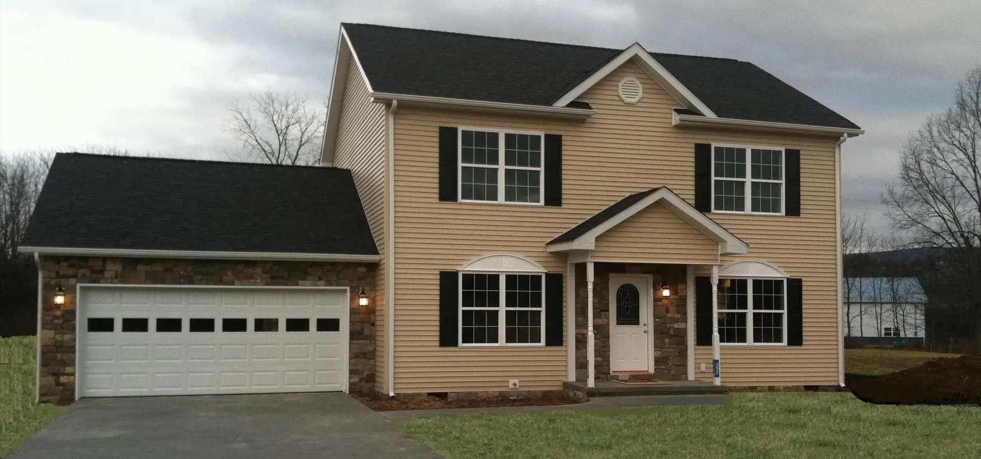 Two-story house with tan siding, black shutters, and attached garage with stone facade.