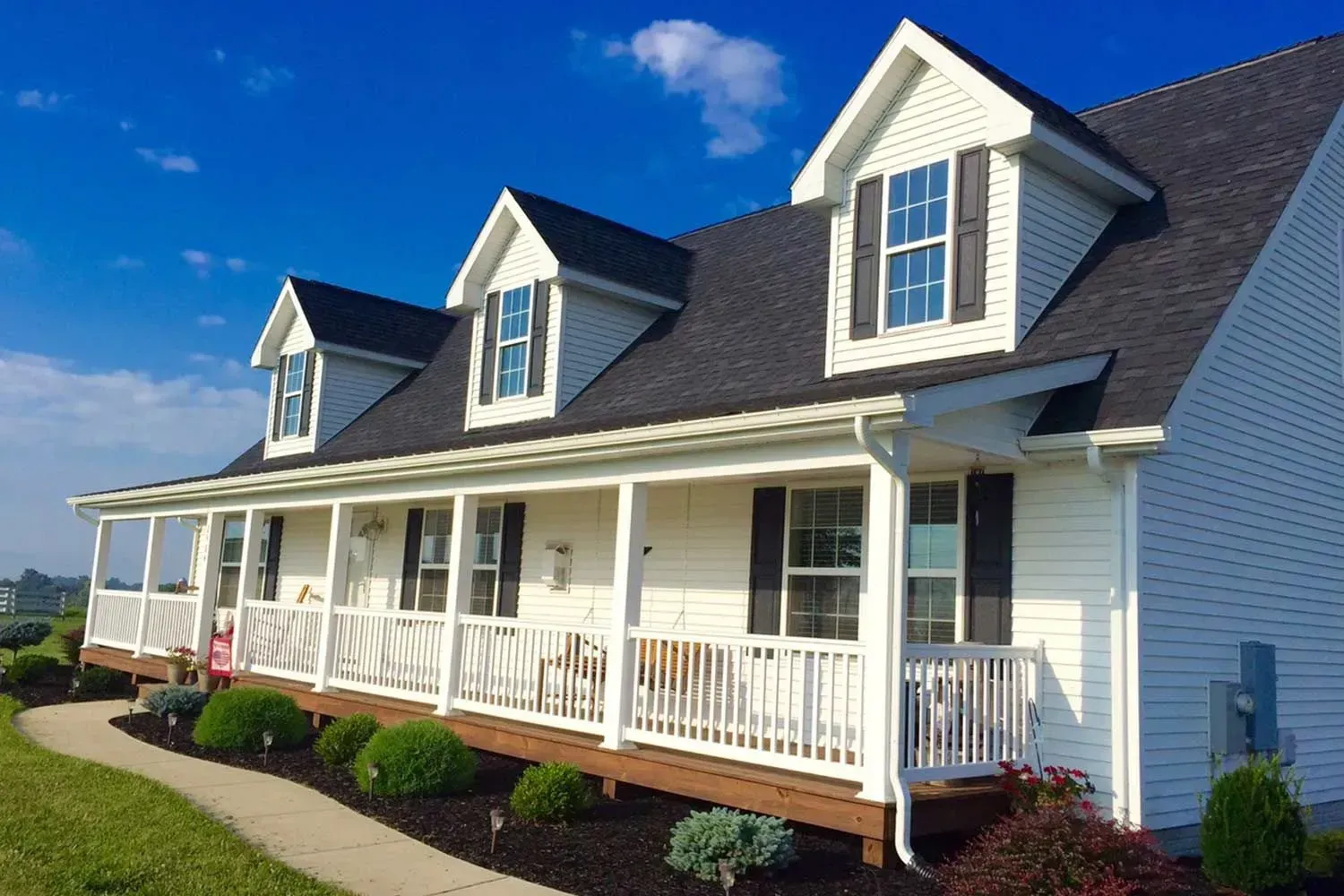 White house with a wrap-around porch, black roof, and dormer windows, on a sunny day.
