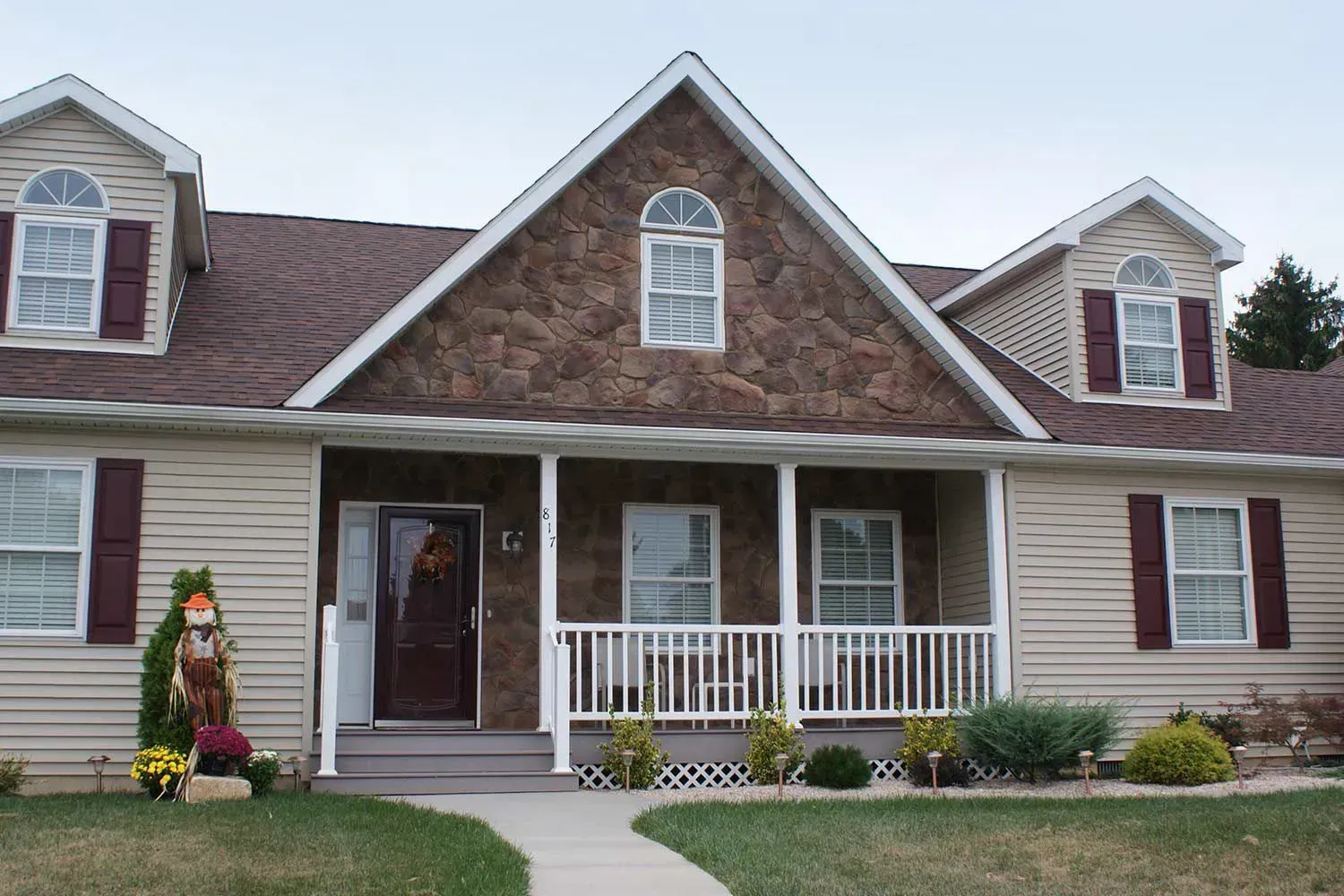 Tan house with brown roof, stone gable, porch with white railing, burgundy shutters, and a walkway.
