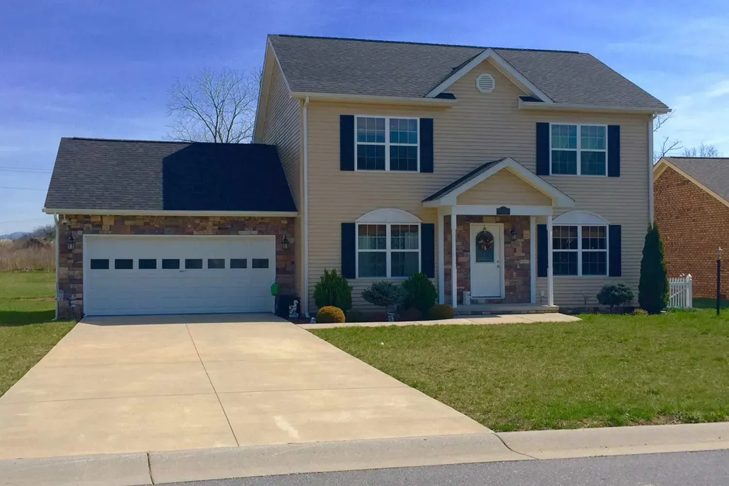 Two-story beige house with a two-car garage, black shutters, and a concrete driveway on a sunny day.