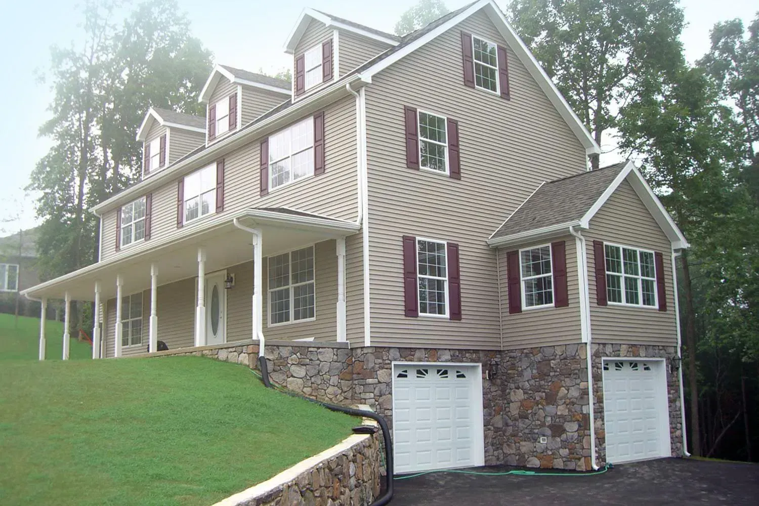 Beige two-story house with a wraparound porch, two-car garage, and stone foundation. Red shutters and dormers.
