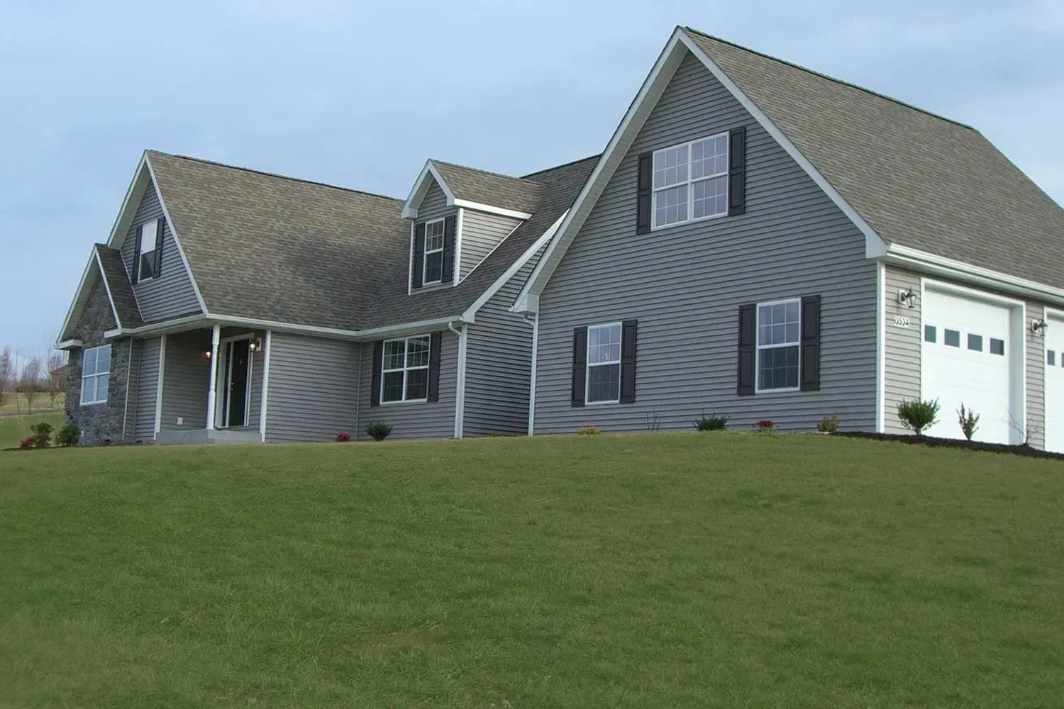 Gray-sided house with a dark roof and white garage doors, set on a green grassy hill.
