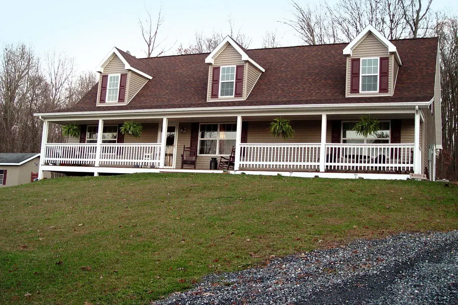 Tan house with porch and dormers, on a grassy hill, with brown roof, white railing and trim.