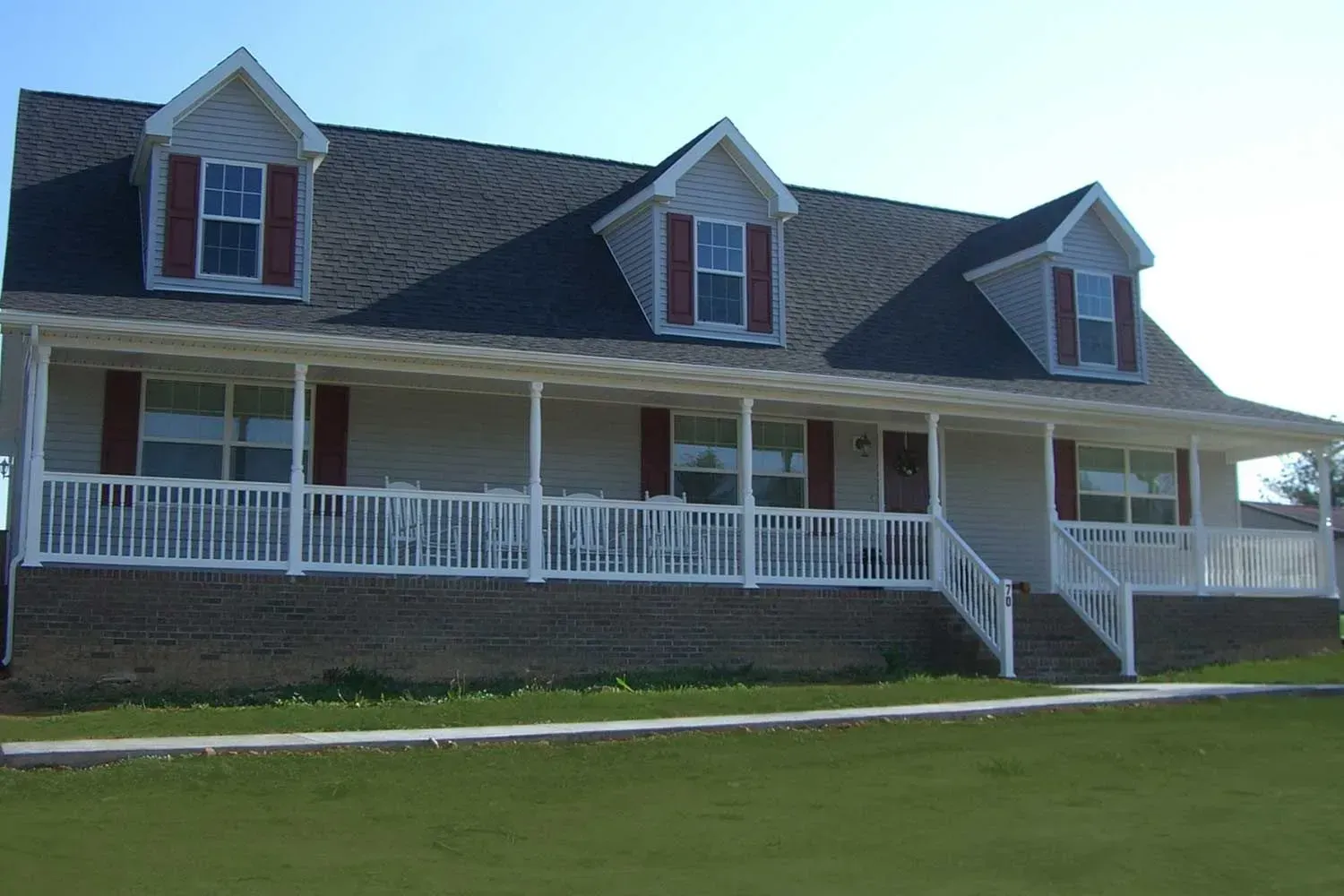 A beige house with a porch, red shutters, and three dormer windows, set on a green lawn.