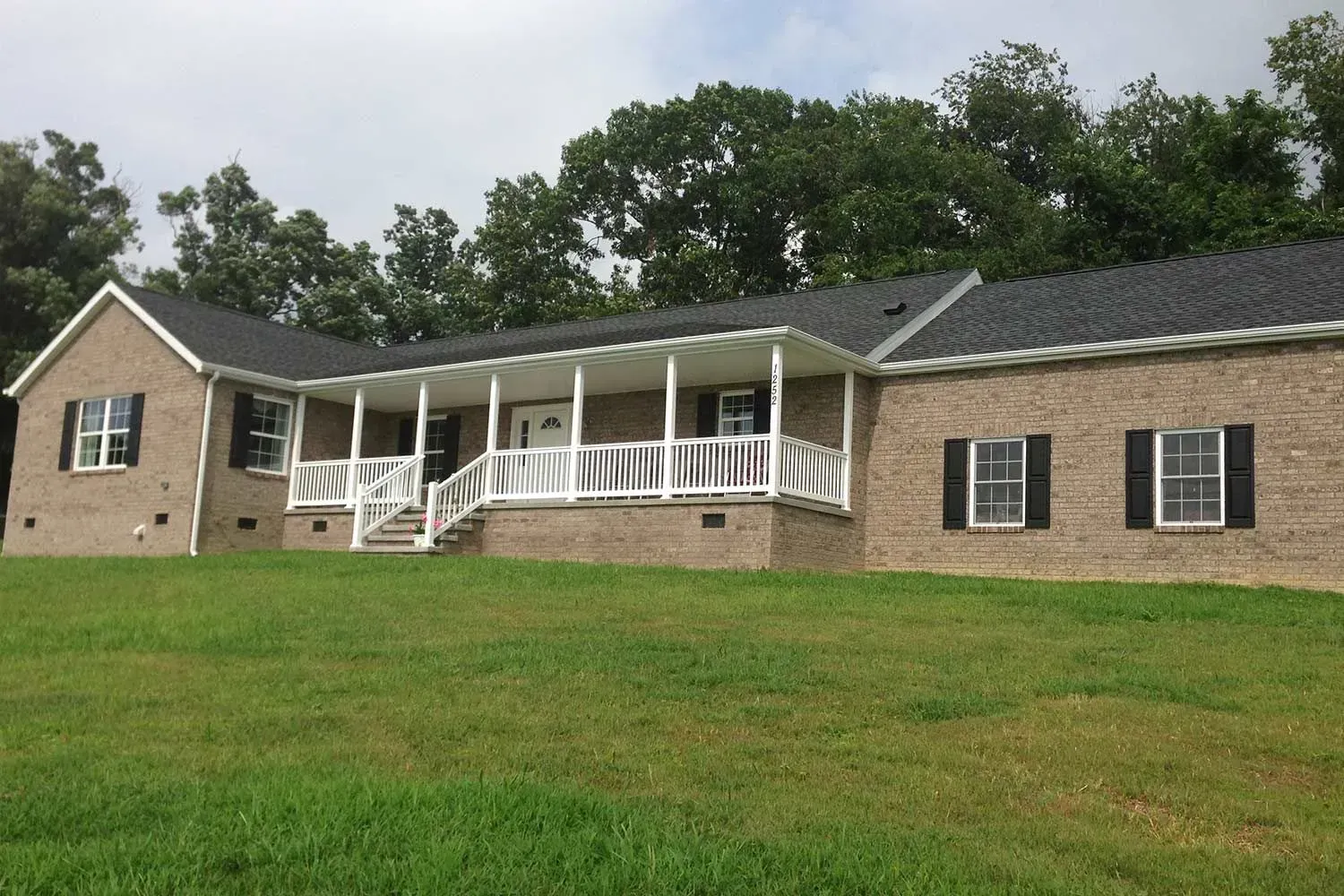 Brick house with a covered porch, white railing, and black shutters on a grassy hill.