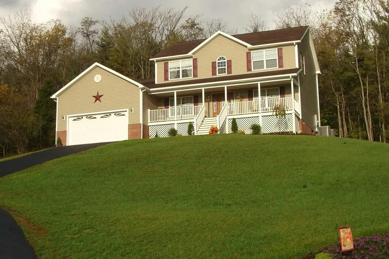 Two-story house with attached garage on a grassy hill. Tan siding, brown roof, and a porch with white railings.