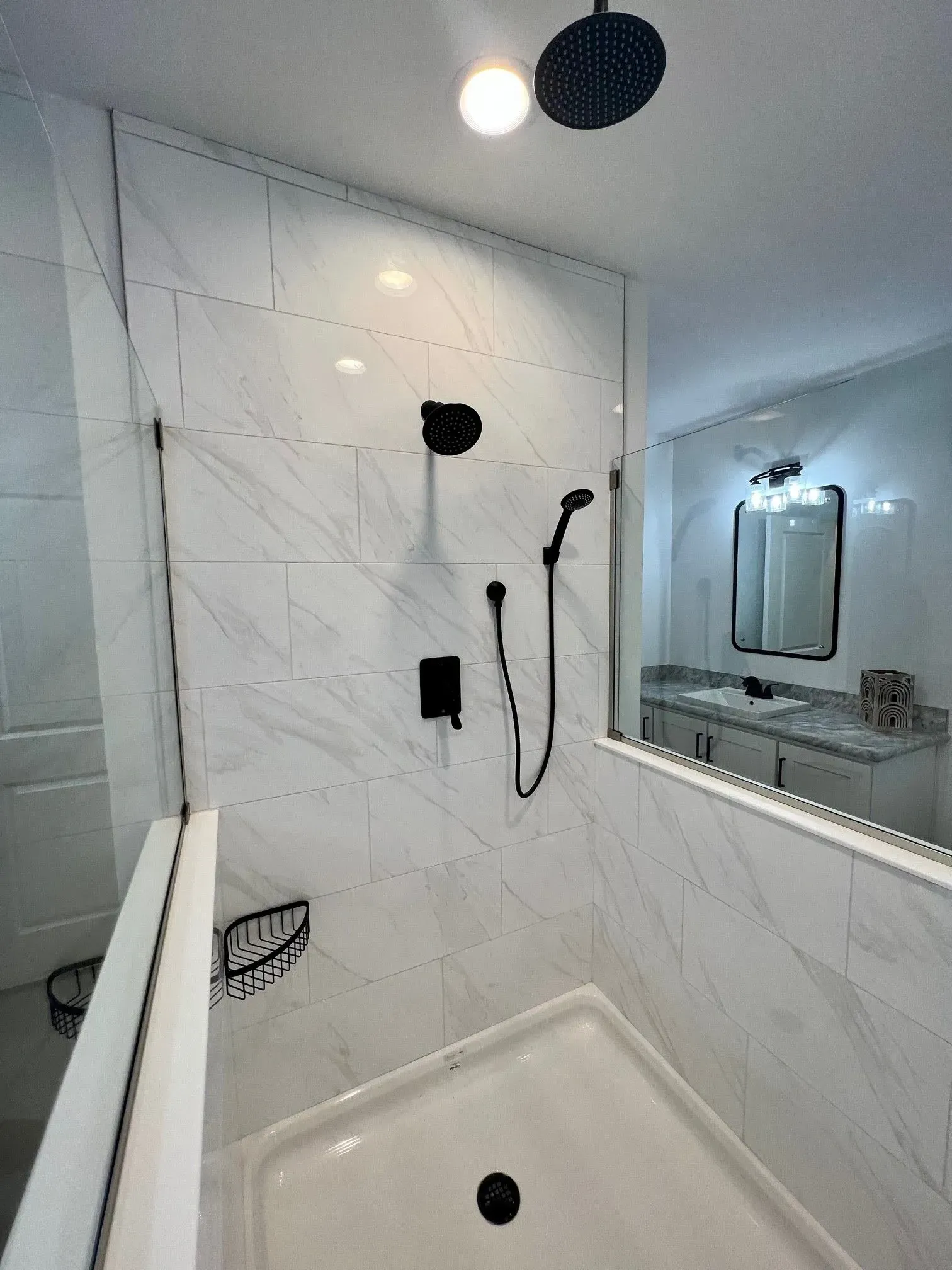 White-tiled shower with black fixtures; showerhead, handheld, and soap dish. Adjacent vanity mirror reflects a double sink.
