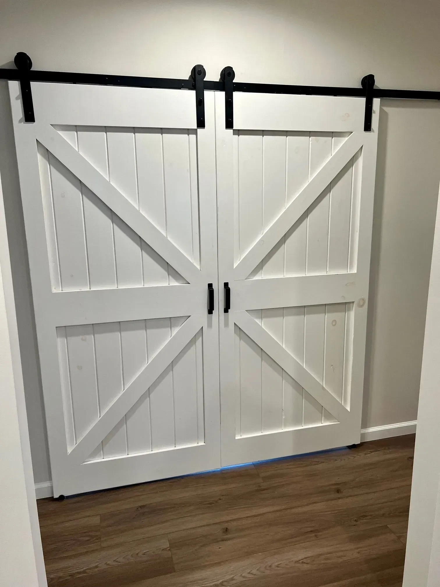 White barn doors with black hardware, spanning a doorway, set against light brown flooring.