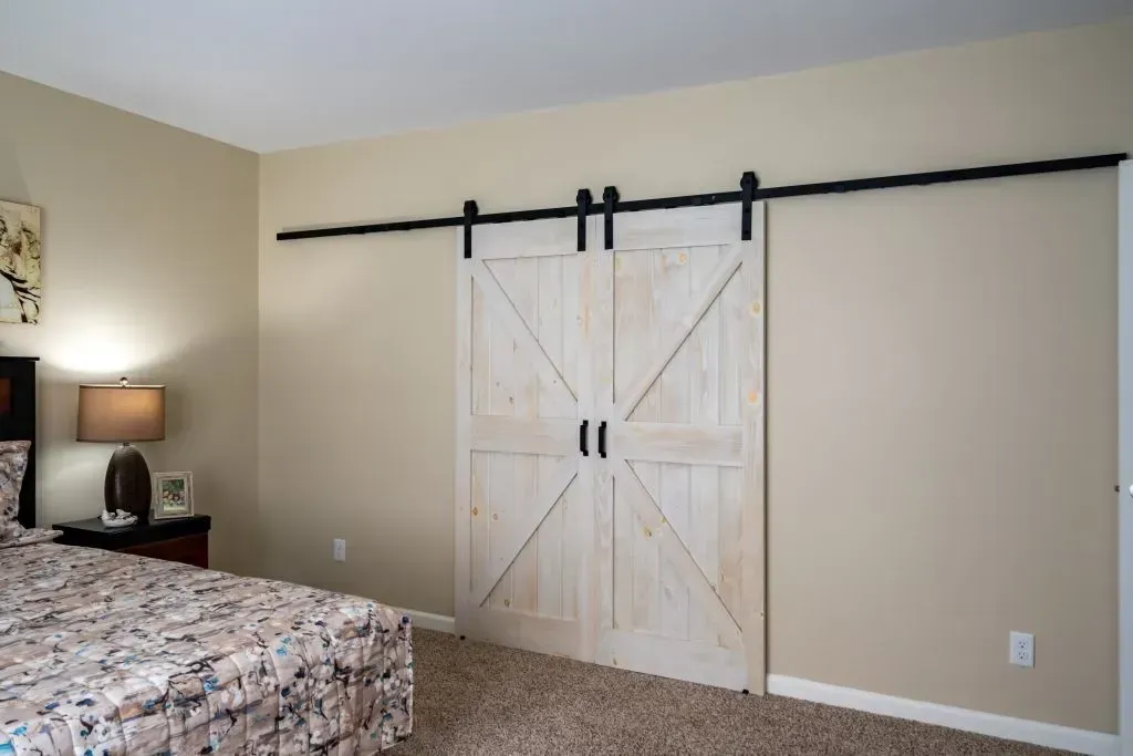 Bedroom with a light-colored wood barn door on a black track against a beige wall, beside a bed with a patterned comforter.
