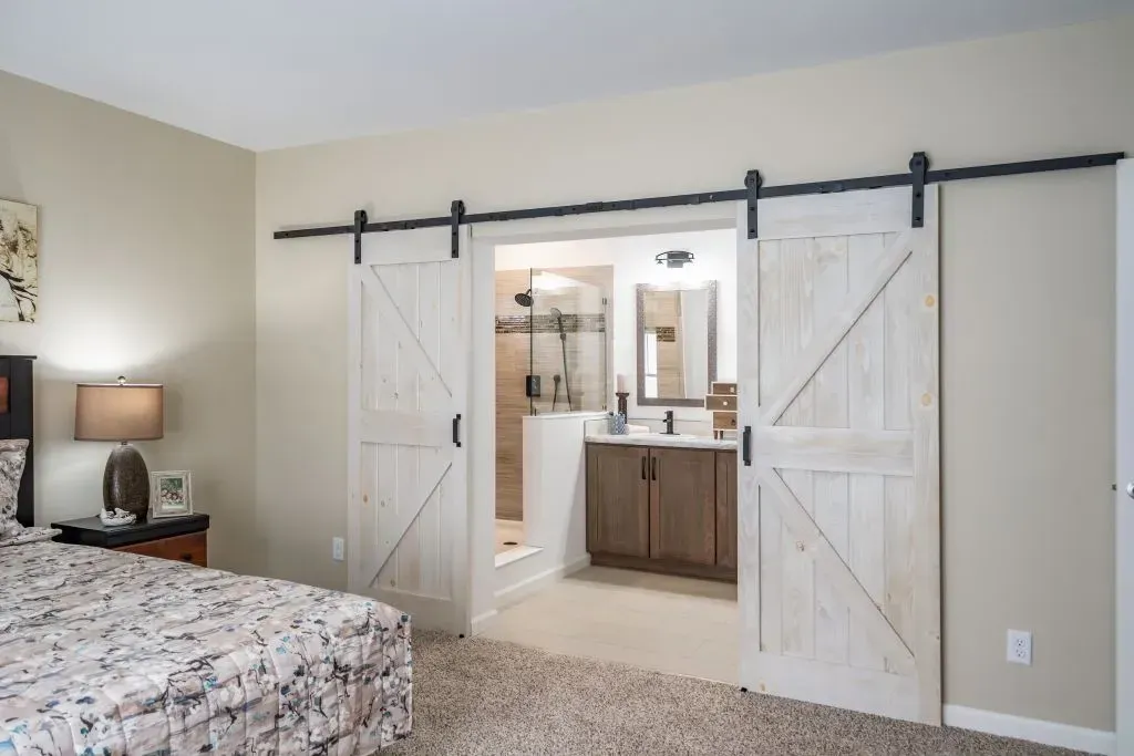 White barn doors lead to a bathroom. Bedroom has a bed, lamp, and neutral carpet.